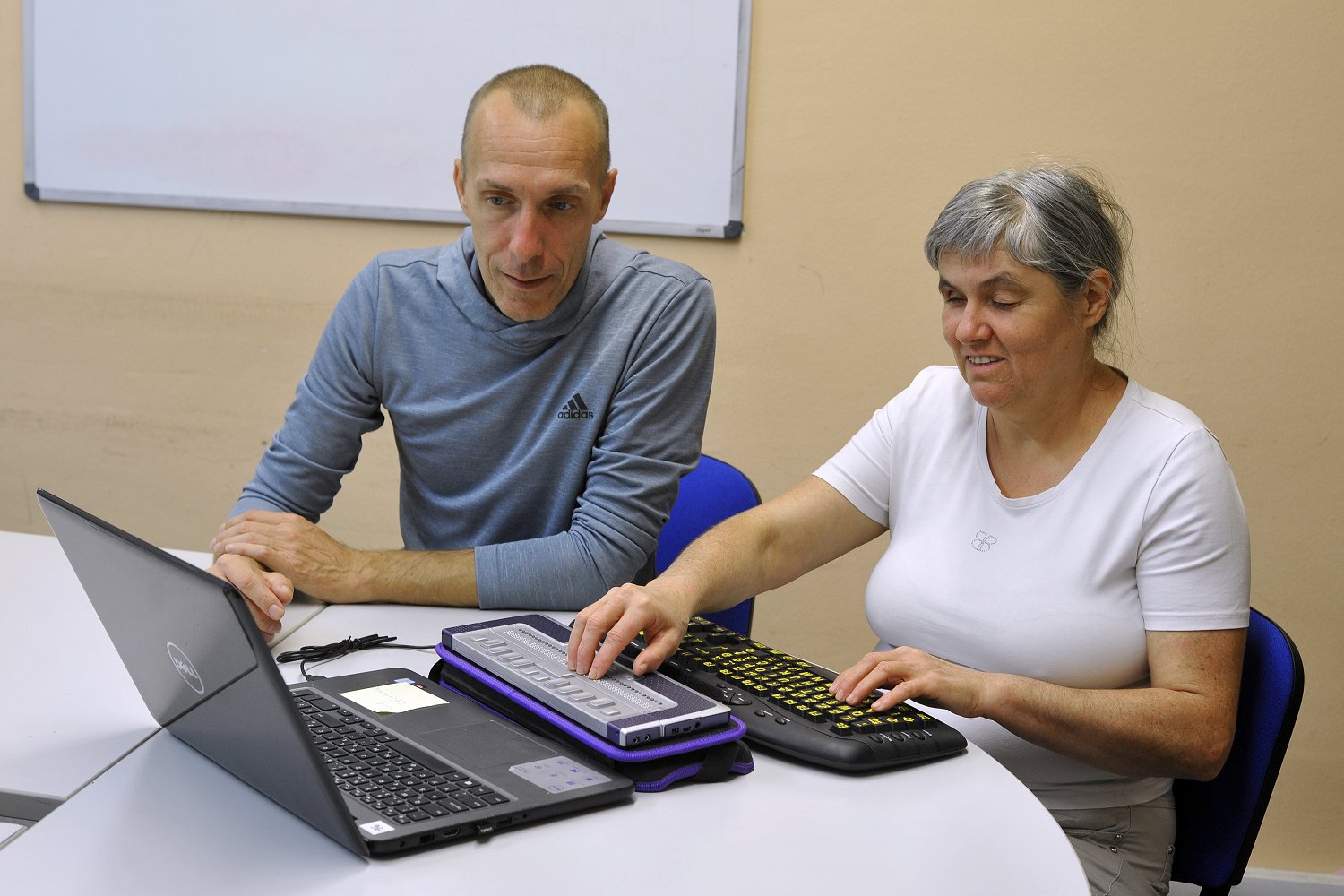 Ženska s slepoto sedi na modrem stolu in se s pomočjo učitelja uči brajeve pisave. Levo roko ima položeno na tipkovnico desno roko na brajevo vrstico. Pred brajevo vrstico je odprt prenosni računalnik črne barve.