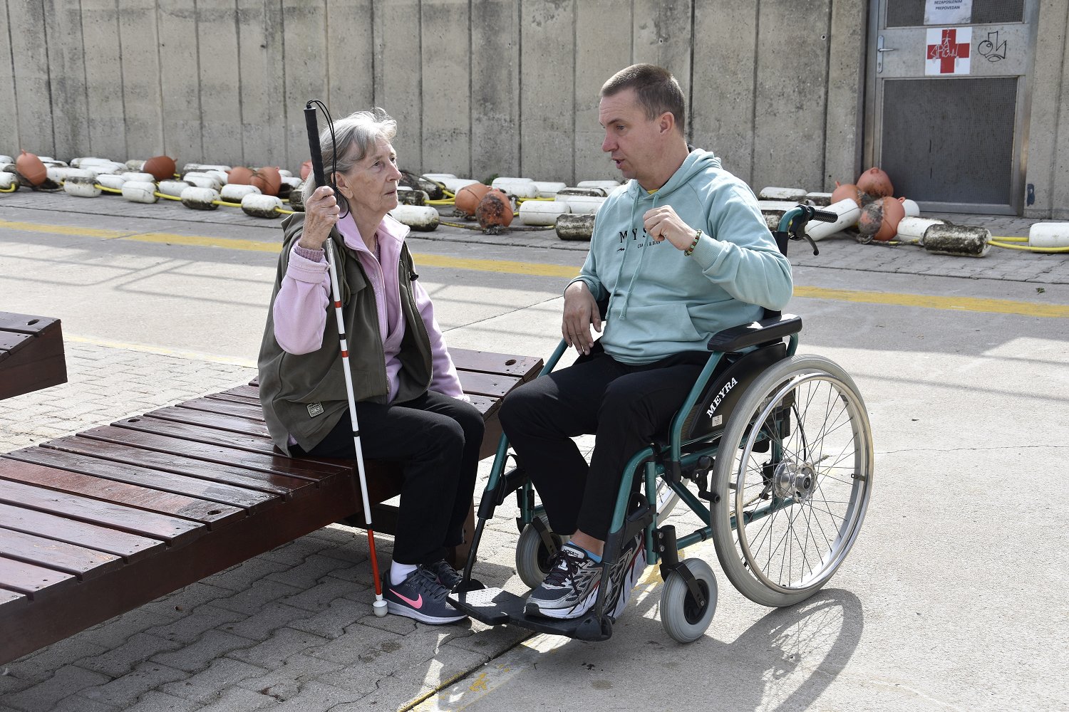 Fotografija prikazuje osebo z belo palico ter osebo na invalidskem vozičku med pogovorom.